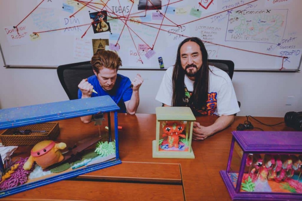 一张 Steve Aoki 和 Seth Green 的照片，他们坐在一张桌子前，手里拿着 Aoki 的 Replicant X NFT 项目的实体头像副本。
